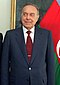 A man in a dark suit with a red tie standing in front of the Azerbaijani flag