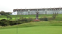 A golf course. In the background is the Turnberry Hotel, a two-story hotel with white façade and a red roof. This picture was taken in Ayrshire, Scotland.