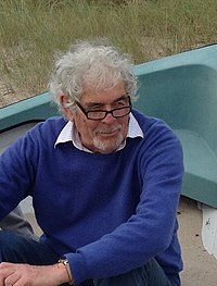 Boulton is sitting on a grassy beach while wearing a blue sweater over a white collared shirt