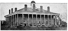 Two-story Greek revival style building with a colonnade surrounding the porch, seven chimneys visible on the roof, and a cuppola at its peak. A group of women are standing in the yard.