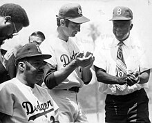 three men wearing baseball caps, two standing and clapping, one seated and looking ahead.