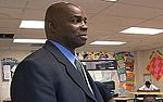 A man, wearing a black jacket with blue shirt, is standing in the middle of a classroom.