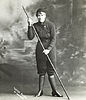 Alice Manfield, or Guide Alice, is pictured in a full length portrait wearing a pant suit consisting of woollen bloomers and a high-necked buttoned jacket pulled in tightly around the waist. She has a type of leggings tightly wrapped around her calves, low-heeled walking boots, a beanie, and is holding a long walking pole.