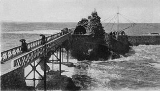 Photographie en noir et blanc d'un rocher sur la mer relié à la côte par une passerelle.