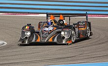 alt=Photographie d'une voiture de sport-prototype gris, noir et orange, vue de trois-quarts, sur une piste.