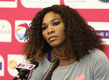 Portrait photographique en couleurs de Serena Williams, l'air sérieux, devant un micro de presse.