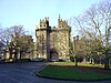 A large stone castle, with imposing towers either side of the gateway, is partly obscured by trees on the green in front of the building. A road leads up to the castle, at the end of which are parked six cars outside the high wooden doors.