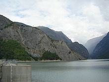 Vue depuis un barrage sur un lac où plonge une crête montagneuse, dans une vallée encaissée.