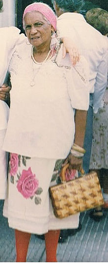 Full-length photograph of a woman in a white blouse with a floral skirt holding a purse.