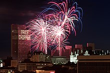 Blue and red fireworks explode over a complex of buildings after dusk.