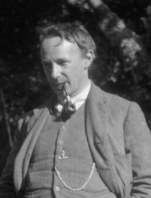 Photographie en noir et blanc d'un homme élégamment vêtu au regard pénétrant, fumant la pipe, une chaîne de montre en évidence sur le gilet de son costume.