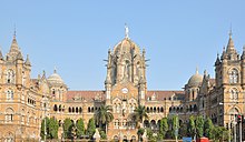 A brown building with clock towers, domes and pyramidal tops. A wide street in front of it