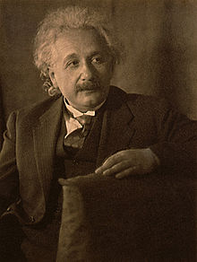 Photographie sépia d'un homme assis aux cheveux gris clair, en coiffure désordonnée, portant une moustache, ayant le regard pensif tourné vers la gauche.