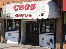 Street view of storefront with doors in the center flanked by windows on both sides. A white awning says "CBGB" in large red letters in a decorative font, and "OMFUG" below that in the same style.