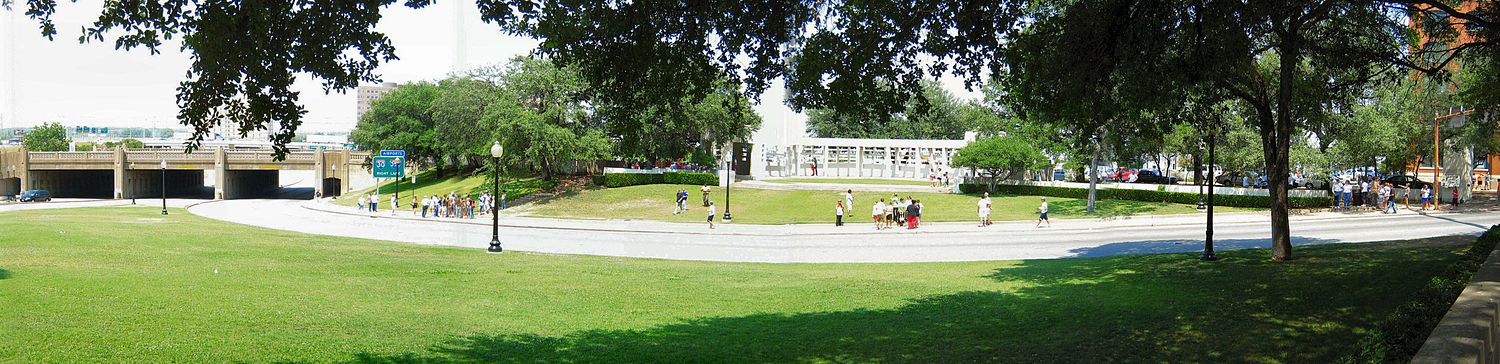 Foto de Dealey Plaza - tute maldekstre troviĝas la fervoja transveturejo, de kie la eventon observis S. M. Holland. Meze estas videbla la herbokovrita monteto kaj blanka parto de betona mureto apartiganta Dealey Plaza de la parkejo; tute dekstre eblas malantaŭ arbokrono vidi parton de la konstruaĵo de Teksasa libra grandmagazeno