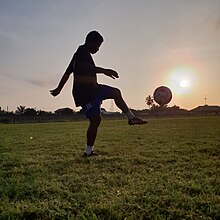 Devant un lever de soleil, dans un pré, une jeune fille fait rebondir un ballon de football sur son pied.