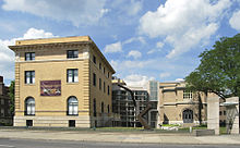 Two classic, buff-colored brick buildings are connected by a modern glass and steel atrium on an urban street.