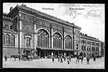 Carte postale montrant partiellement et en perspective la gare de Strasbourg, un imposant bâtiment de style wilhelmien, avec son entrée par la partie centrale et son aile droite. Ses façades, librement inspirées du style Renaissance, s'ouvrent par de grandes baies vitrées. Devant l'auvent de la gare stationnent plusieurs personnes et chevaux attelés ou montés. Les impressions en allemand Strassburg et Zentralbahnhoff figurent au-dessus de la gare parmi d'autres mots manuscrits.