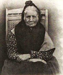 An elderly woman sitting in a high-backed wooden chair wearing a shawl partly covering her head with her arms crossed in her lap.