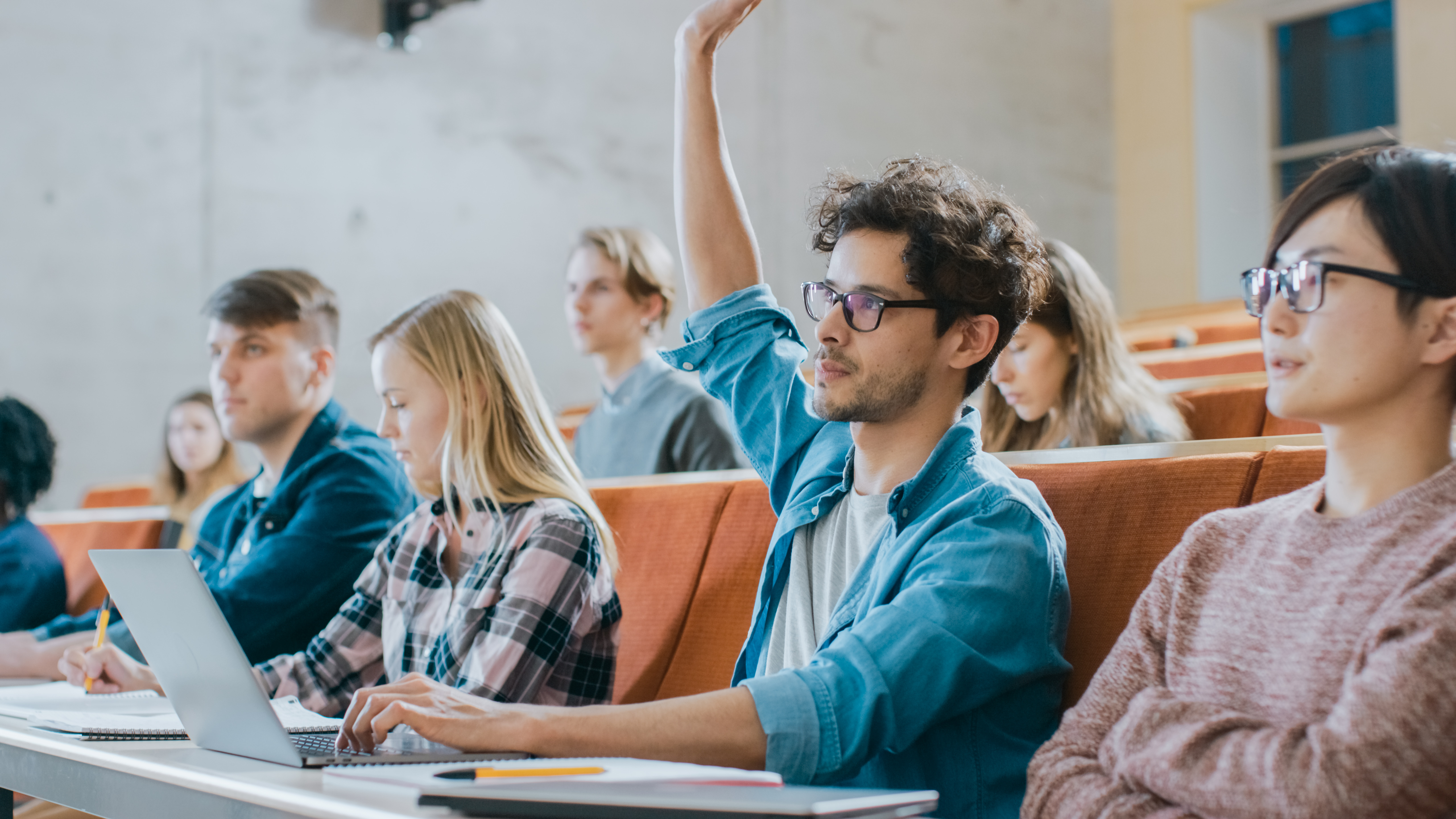 An auditorium of students in a college class. Photo is focused on one male student raising their hand