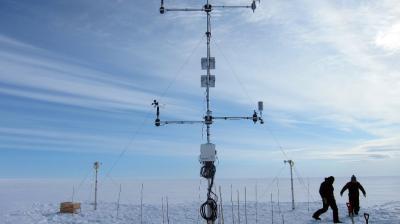 Two scientists working on a complex weather station apparatus on a snow-covered plain under a clear blue sky.