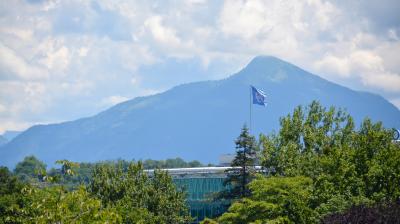 A glass building is surrounded by greenery with a flag flying in front of it. In the background, there is a mountain range under a partly cloudy sky.