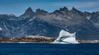 A large iceberg floats in a calm blue sea with rocky mountains in the background under a clear sky.