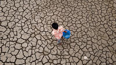 A person carrying a blue bucket walks on cracked, dry earth, suggesting a drought or water scarcity.