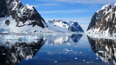 A serene icy landscape shows snow-covered mountains reflecting in the calm waters of a narrow waterway, with floating ice chunks scattered throughout.