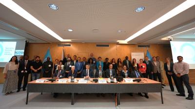 A group of individuals stand behind a conference table with microphones and nameplates in a formal meeting room. Participants are dressed in business attire.