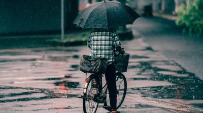 Person riding a bicycle in the rain, holding a large black umbrella and carrying two bags on the bike. The street is wet with visible puddles.