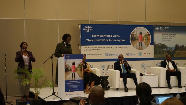 Panel discussion at an event featuring the UN Global Early Warning Initiative for climate adaptation. Four panelists are seated on stage, while two people stand at podiums with a presentation screen in the background.