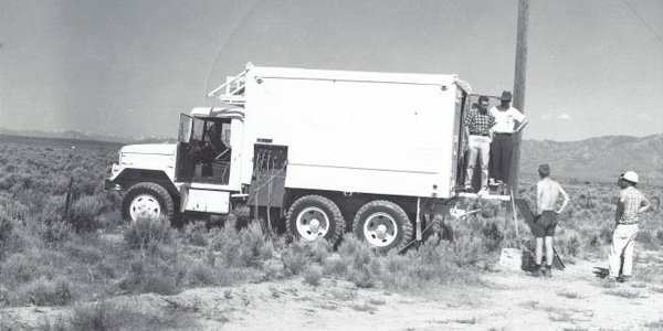 box truck parked on scrublands with four men standing in the back of the truck and on the ground behind the truck. There is a tower behind them.