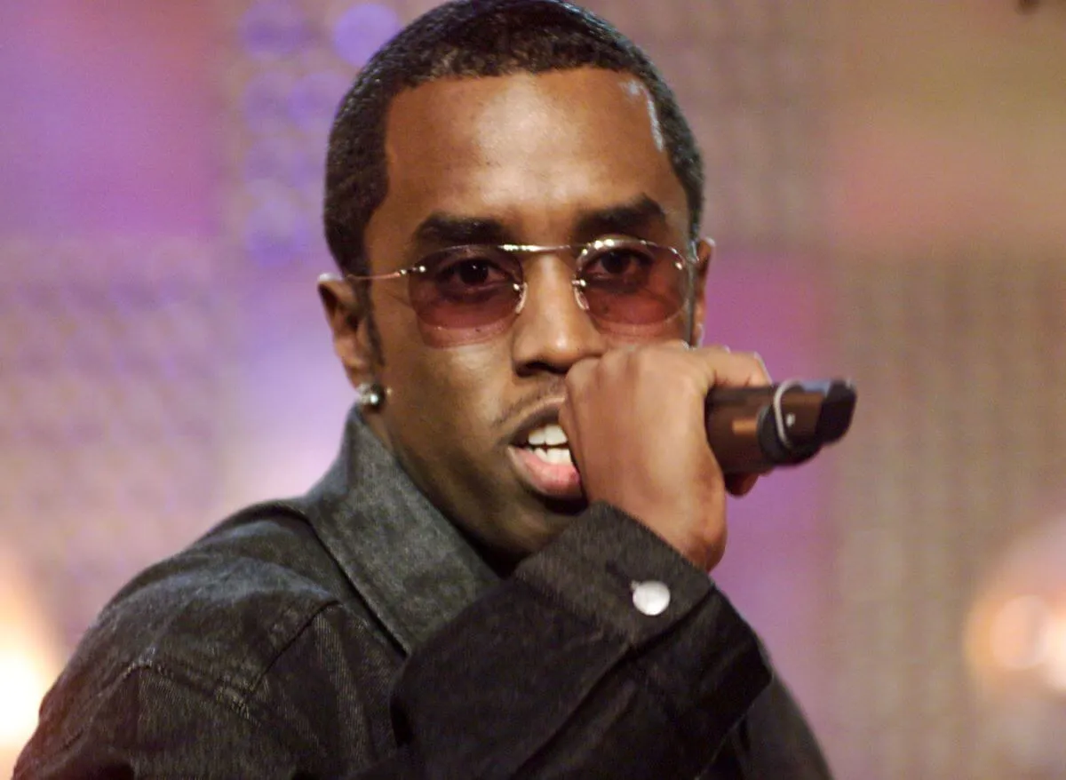 Sean 'Diddy' Combs wears a black denim jacket and sunglasses. He holds a microphone.