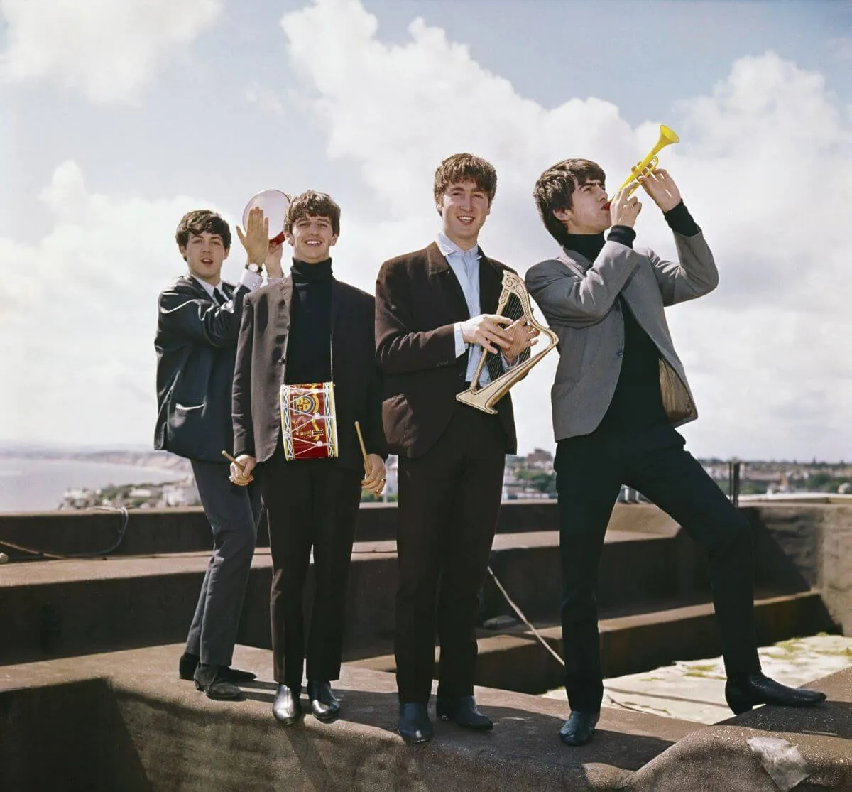 The Beatles stand on a rooftop and hold small instruments.