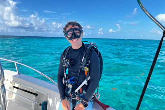 Rob Harper stands on a boat in diving gear in the middle of vibrant blue waters