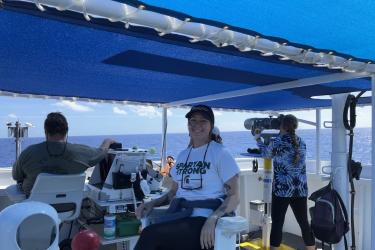 Micahela sits on a chair aboard a ship under a blue tent while two people observe the ocean on each side of her. 