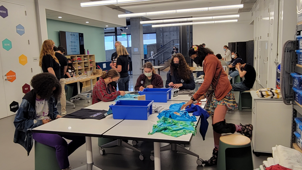 Teens collaborate around a table to work on an activity to upcycle used shirts. There are other groups of people behind them working on different activities at various tables. 