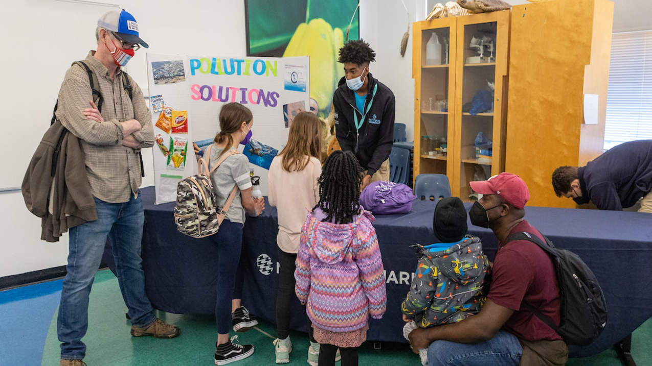 A person is standing behind a long table inside a room and is presenting to six people using a poster board that is set up to the person’s left that reads “Pollution Solutions.” There is a cabinet behind the person and another person crouching down behind the table to the right.