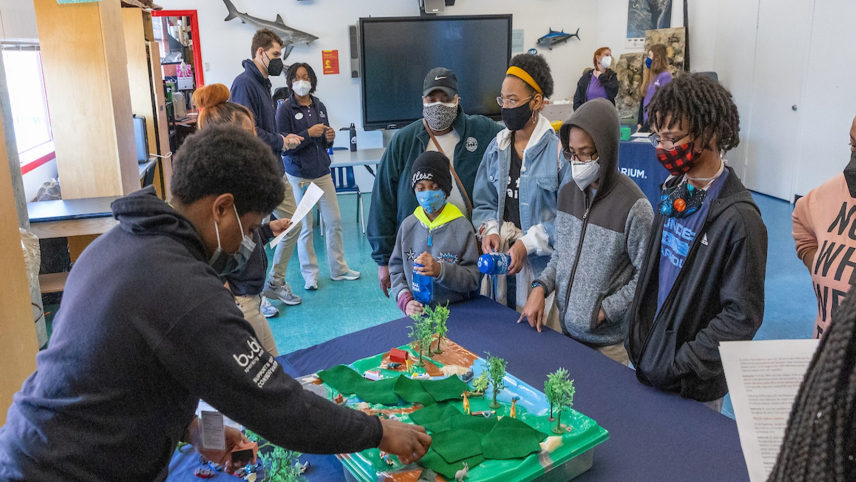 A person is standing behind a table and using a display that looks like a plastic landscape with toy trees, boats, people, and green felt for grass to engage with six people standing in front of the table. There are five other people standing around in the room.