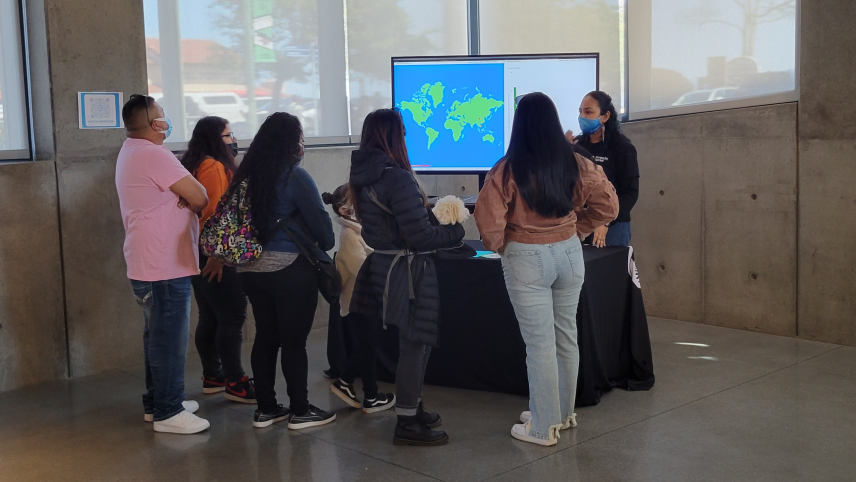 Six people stand in front of a table listening to another person behind the table talk. There is also a TV screen behind the table displaying a map of the world. 