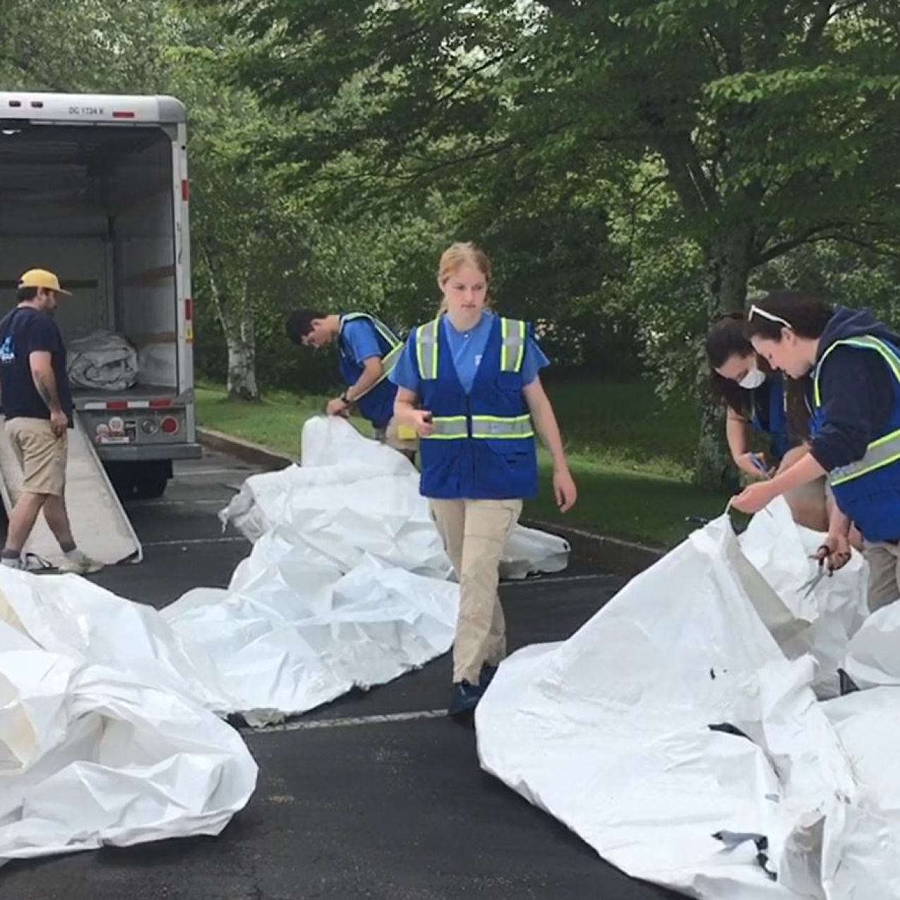 Six people are working outside in a parking lot next to a line of trees. Three people are holding onto large pieces of shrink wrap that are laid out on the parking lot. There is a truck parked behind the group of people with a ramp for loading.