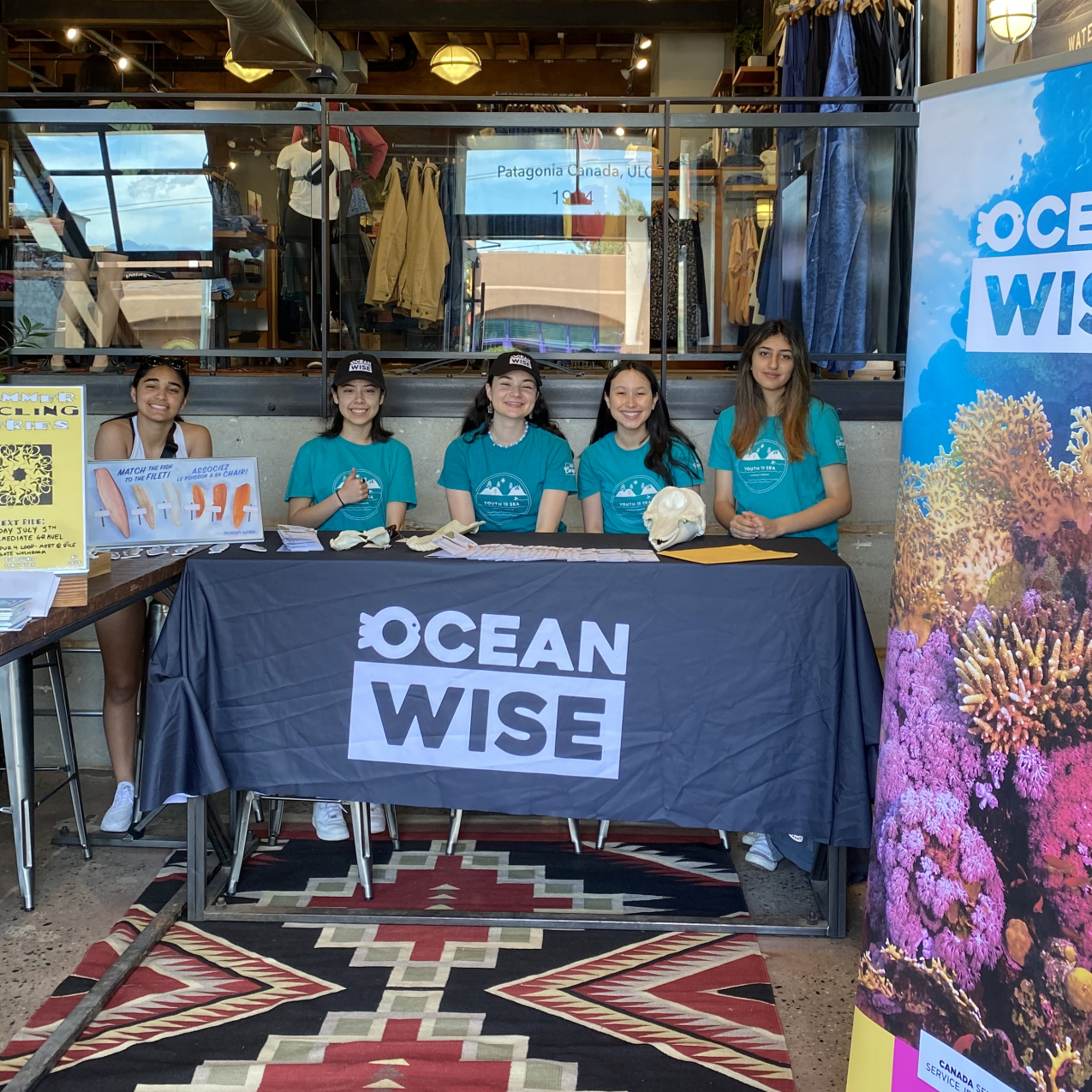 Five people sit behind a table that is draped with a navy blue table cloth that reads “Ocean Wise.” Various papers and animal bones are displayed on the table. To the left is another table with multiple signs. To the right of the table is a long banner with an underwater photo of colorful coral that reads “Ocean Wise.” In the background, there is railing covered by glass and clothes on display for sale. 