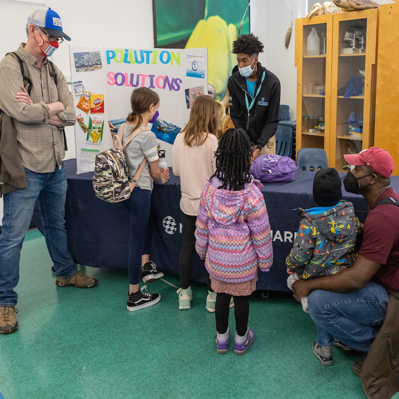 A person is standing behind a long table inside a room and is presenting to six people using a poster board that is set up to the person’s left that reads “Pollution Solutions.” There is a cabinet behind the person and another person crouching down behind the table to the right.