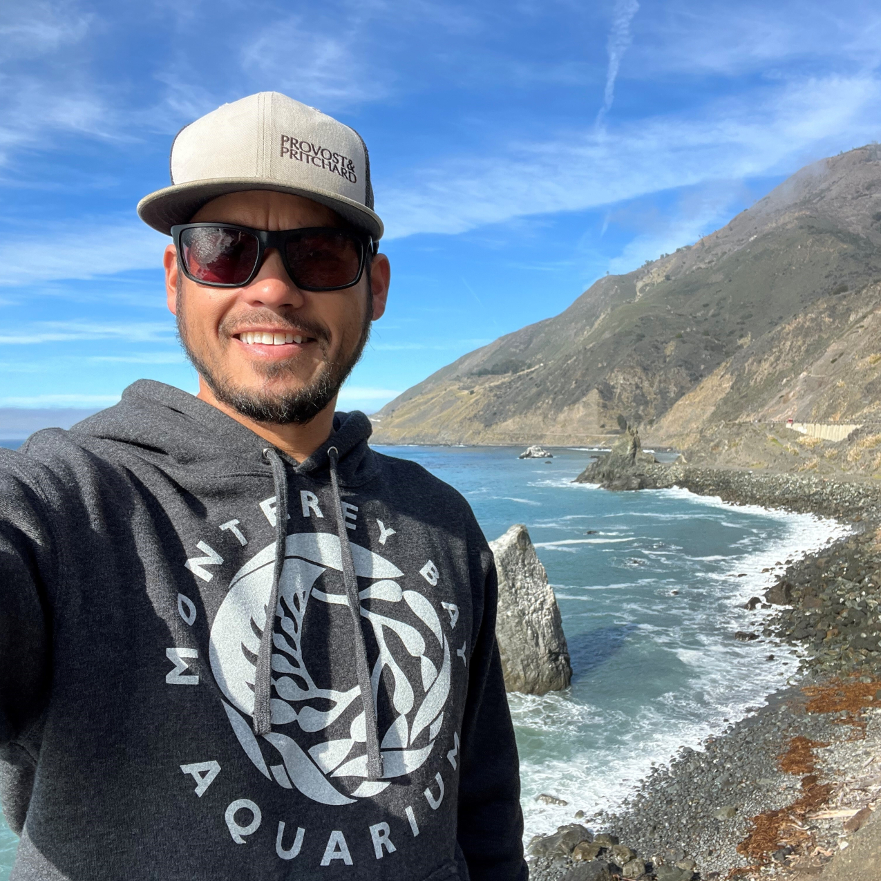 Philip Melcher, wearing a hat and sunglasses, is standing in front of a beach on the California coastline, with large mountains in the background. 