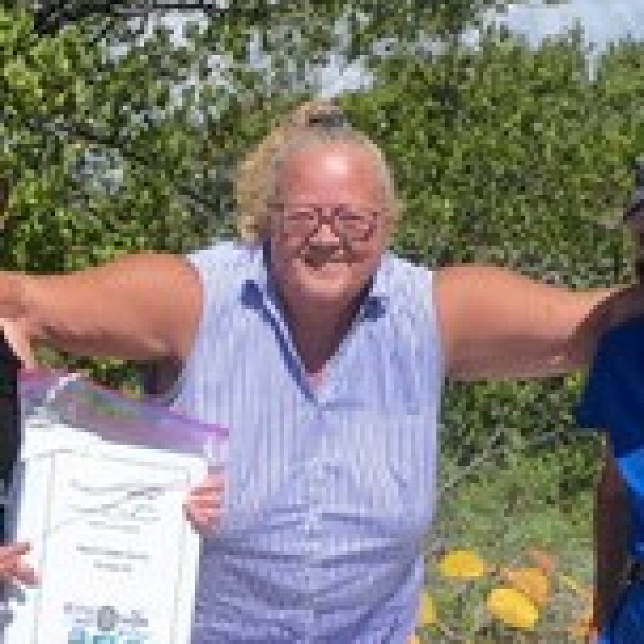 Linda Palewicz standing on a beach, with shrubbery behind her, in front of materials necessary for horseshoe crab tagging. Her arms are outstretched around the shoulders of two people next to her who are not show in the picture.