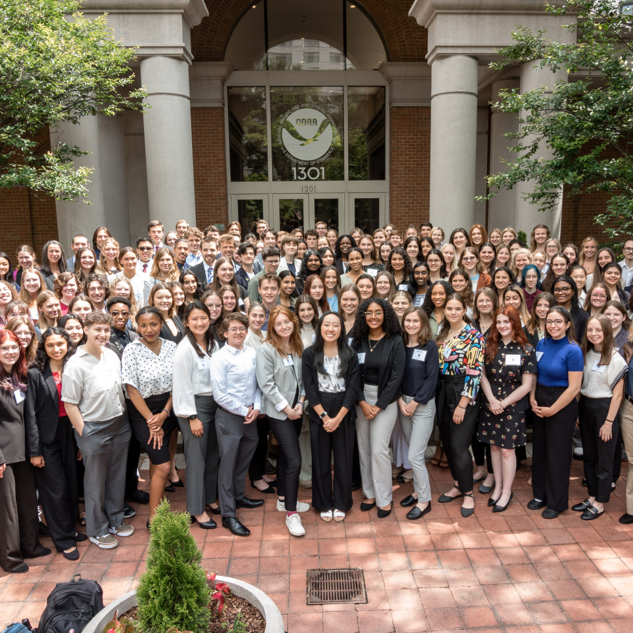 A group approximately 130 people in business casual attire pose together outside of a large brick building that has a NOAA logo above the door.