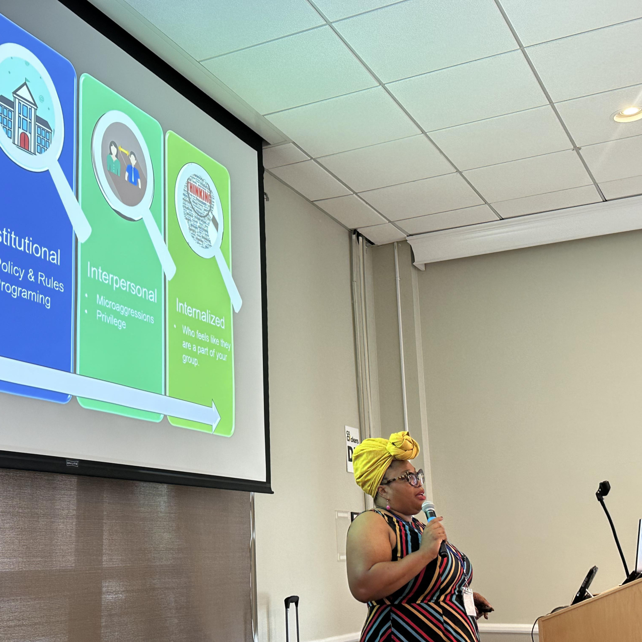 A woman in a yellow headwrap and a dress stands with a microphone in her hand.