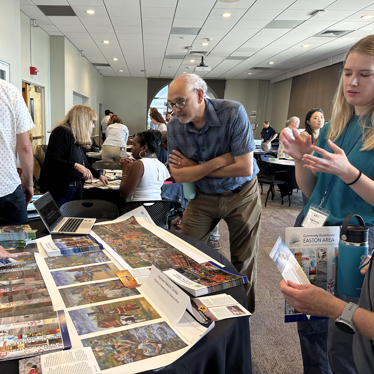 A woman in a green shirt talks to others standing over a table covered with a blue-table cloth. On the table are colorful models of murals, booklets, and other flyers.