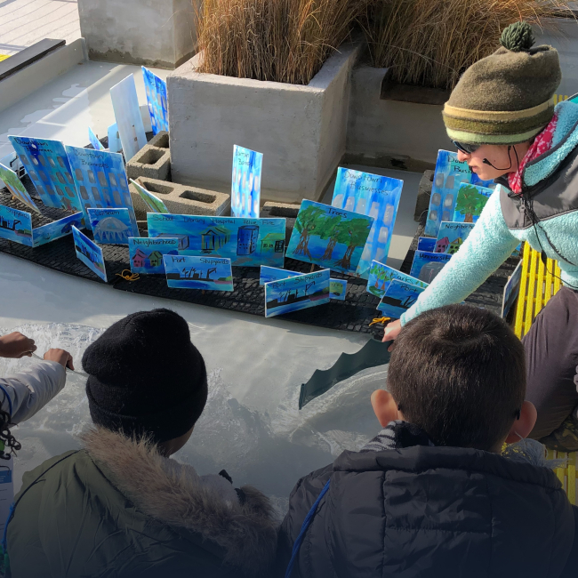 Three students use pieces of plastic and board to push water towards a replica of Norfolk. An instructor assists the students with the activity.